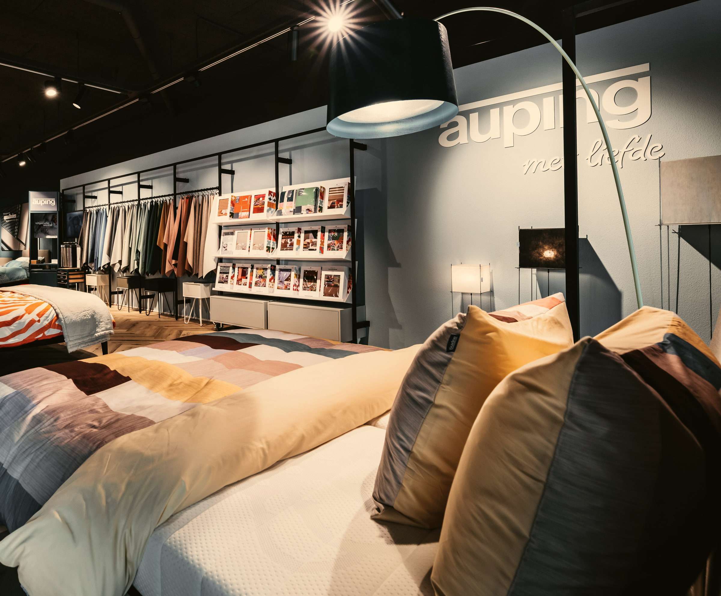 De Auping Studio van Wielinga & Tabbers: een opgemaakt bed met kleurige kussens onder een booglamp, voor een blauwgrijze wand met het Auping-logo en een rek met bedtextiel